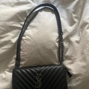Svart YSL quiltad axelväska i skinn (ej äkta) - Snygg svart axelväska i quiltat skinn med silverfärgad  detaljer. Den är aldrig använd då jag fick den i present, så super bra skick. Den går och ha på olika sett då man kanhända själv vilket band man vill ha, om man vill har kortare eller längre. Den är perfekt till både fest och vardag.