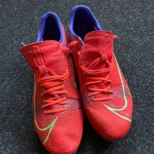 Nike Mercurial röda fotbollsskor - Säljer ett par Nike Mercurial fotbollsskor i rött med gröna och blå detaljer. Använda någon säsong på gräs så knappt slitna nästan ny skick. Pris kan diskuteras. Skorna har snörning, låg profil och mönstrad ovandel för extra grepp. Sulan är silverfärgad med röda och gröna dobbar. Perfekta för dig som vill sticka ut på planen.