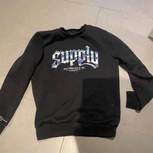 Svart sweatshirt Supply & Demand - Svart sweatshirt från Supply & Demand med stort blått och vitt tryck 'supply' på bröstet i coolt typsnitt. Liten vit text på ena ärmen. Tröjan har rund hals, ribbade muddar och är gjord i mjuk bomullsmix. Perfekt för en streetwear-look.