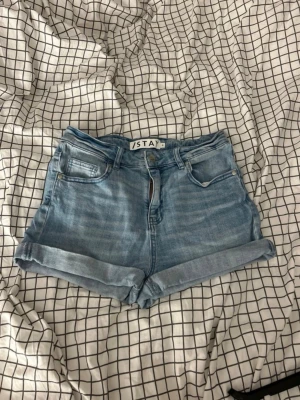 Blå denimshorts från Stay - Säljer ett par klassiska blå denimshorts från Stay med upprullade benslut och femficksmodell. Shortsen har normal passform, bälteshällor och dragkedja framtill. Perfekta till sommaren och lätta att matcha med allt.
