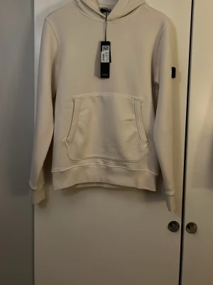 Vit hoodie från stone island  - Riktigt snygg vit hoodie från stone island. Den har den klassiska badgen på vänstra armen och en fika på magen. Den har även en luva. Fick den i julklapp men den var fel storlek och är därför aldrig använd och har alla tags och lappar kvar. Hör av er vid frågor! 