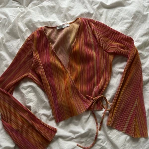 Glittrig omlottopp från Costes Festival - Säljer en färgstark omlottopp från Costes Festival med långa utsvängda ärmar. Toppen har smala ränder i rosa, orange, gul och glitter. Knytband i midjan och v-ringad skärning. Perfekt för dig som vill sticka ut med en unik och trendig look.