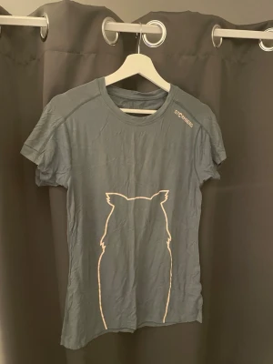 Grå t-shirt med kattmotiv från Stormberg - Snygg grå t-shirt från Stormberg med korta ärmar och ett stilrent kattmotiv i vitt på framsidan. T-shirten har rund halsringning och är gjord i mjuk bomull, perfekt för en avslappnad look. Stormberg-loggan syns på axeln.