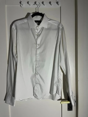 Vit skjorta Riley Slim Fit - Klassisk vit skjorta från Riley i slim fit-modell. Skjortan har långa ärmar, knappar framtill och en stilren krage. Tillverkad i mjuk bomull som känns skön mot huden. Perfekt för dig som gillar en clean och enkel look.