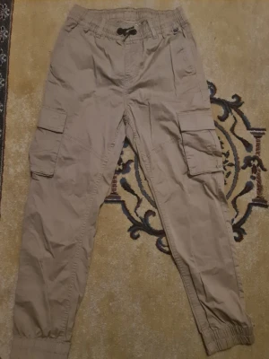 Beiga cargobyxor KappAhl strl 104 - Säljer ett par beiga cargobyxor från KappAhl i storlek 104. Byxorna har resår i midjan med snörning, stora sidofickor med lock och muddar vid bensluten. Perfekta för en avslappnad och cool stil.