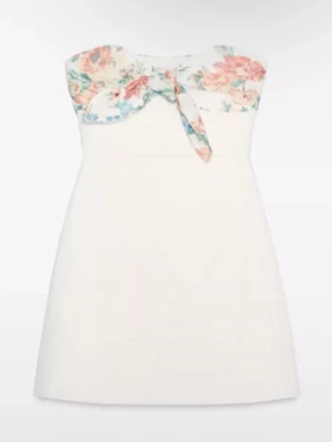 Vit kort klänning med blommig rosett - Supersöt vit kort klänning med axelbandslös design och en stor blommig rosett framtill. Överdelen har ett blommigt mönster i pastellfärger och knyt i mitten. Kjolen är enkel och A-linjeformad. Perfekt för sommarens alla tillfällen!