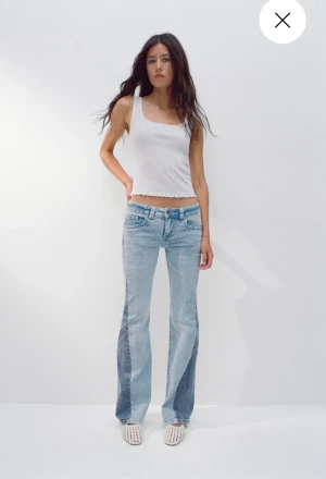 Ljusblå bootcut jeans med mörka paneler - Säljer ett par ljusblå bootcut jeans med coola mörkblå paneler längs sidorna. Jeansen har låg midja, klassiska fickor och är tillverkade i denim. Perfekta för dig som vill sticka ut med en unik look.