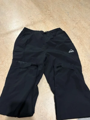 Svarta täckbyxor från McKinley S - Svarta täckbyxor från McKinley med elastisk midja och stora fickor med kardborrestängning på benen. Perfekta för utomhusaktiviteter under kyligare dagar. Tillverkade i slitstarkt syntetmaterial som står emot vind och väta.