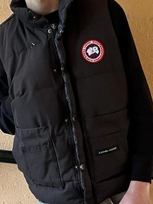 Svart dunväst från Canada Goose - Säljer en svart dunväst från Canada Goose med hög krage och stora framfickor. Västen har tryckknappar och dragkedja framtill samt den klassiska Canada Goose-loggan på bröstet. Perfekt för lager på lager under kyliga dagar.