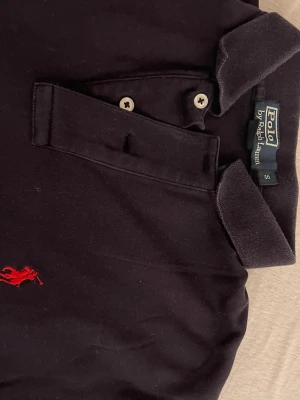 Svart piké från Polo Ralph Lauren - Klassisk svart pikétröja från Polo Ralph Lauren med röd broderad logga på bröstet. Tröjan har korta ärmar, krage och två knappar framtill. Tillverkad i mjuk bomull som känns skön mot huden.