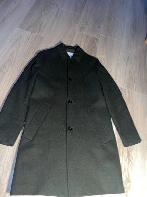 Svart ullrock från BLÄCK - Klassisk svart rock från BLÄCK i ullmix med rak passform och stilrena detaljer. Jackan har krage, knäppning med tre knappar framtill och två snedställda fickor. Perfekt för dig som vill ha en clean och tidlös look.