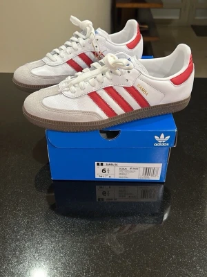 Adidas Samba OG vit/röd sneakers - Adidas Samba OG sneakers i vitt skinn med klassiska röda stripes och detaljer i beige mocka. Skorna har brun gummisula, rund tå och snörning. Perfekta för dig som gillar retrovibbar och tidlös streetstyle.