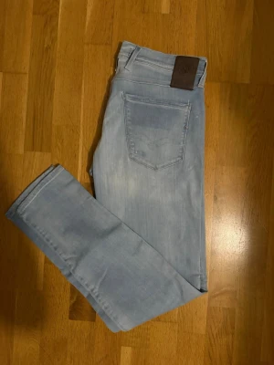 Ljusblå Replay jeans Anbass hyperflex  - Säljer ett par ljusblå jeans från Replay med klassisk femficksdesign och snygga slitningar. Jeansen har raka ben och slim passform, samt Replay-logga.