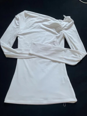 Vit oneshoulder långärmad topp - Säljer en stilren vit långärmad topp med oneshoulder-design och lätt rynkad detalj vid axeln. Toppen är figurnära och gjord i ett mjukt, stretchigt material som känns skönt mot huden. Perfekt att matcha med jeans eller kjol för en trendig look.