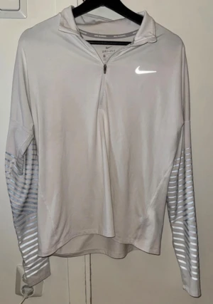 Vit Nike långärmad träningströja - Nike Dri-Fit långärmad träningströja i vit med gråa ränder på ärmarna. Tröjan har half zip framtill och ett reflekterande Nike-logga på bröstet. Perfekt för löpning eller gym, med lätt och ventilerande polyester-material.