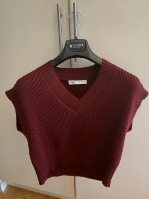 Vinröd stickad väst från Zara - Säljer en vinröd stickad väst från Zara med v-ringad hals och ribbad struktur. Modellen är croppad och har breda axelpartier utan ärmar. Perfekt att styla över en skjorta eller t-shirt för en trendig look.