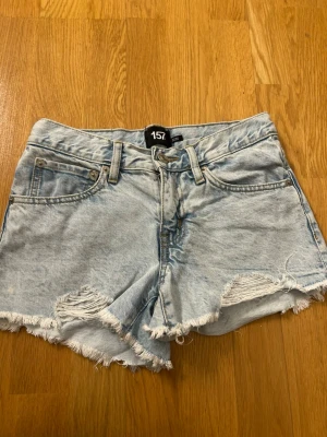 Ljusblå denimshorts från 157 - Säljer ett par ljusblå denimshorts från 157 med råa, fransiga kanter och slitna detaljer framtill. Klassisk femficksmodell med bälteshällor och knappgylf. Perfekta för varma dagar och en avslappnad look.