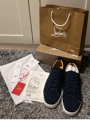 Mörkblå Christian Louboutin sneakers - Säljer ett par mörkblå sneakers från Christian Louboutin med klassisk röd sula och svarta nitar på tån. Skorna har snörning, rund tå och vit platt sula. Tillverkade i skinn och textil, med exklusiv design och detaljer. Kommer med originalkartong, dustbag och äkthetsbevis.