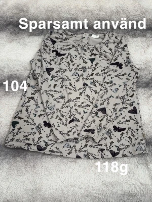 Blommig långärmad topp med fjärilar - Söt långärmad topp i grå med blommigt mönster och fjärilar. Rund halsringning och mjukt, stretchigt material som känns skönt mot huden. Perfekt för dig som gillar naturinspirerade prints och vill ha en bekväm vardagstopp.🌸 Sparsamt använd. Storlek 104