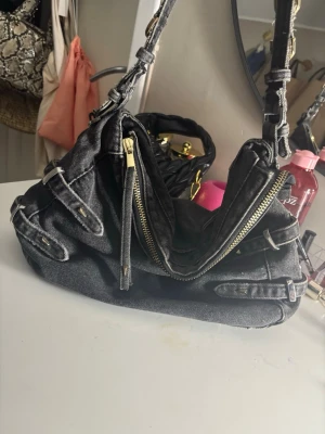 Svart denim axelväska med detaljer - Cool svart axelväska i denim med flera spännen och dragkedja i guld från Zara. Väskan har en unik form och justerbar axelrem med metallringar. Perfekt för dig som gillar streetstyle och vill ha en väska som sticker ut. Har använts 3 gånger 