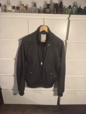 Mörkblå bomberliknande jacka från J.Lindeberg - Säljer en Marinblå bomberjacka från J.Lindeberg med klassisk ståkrage och dragkedja framtill. Jackan har två fickor med knappstängning och ribbade muddar vid ärmslut och midja. Enkel och stilren design som funkar till det mesta. Strl S-XS