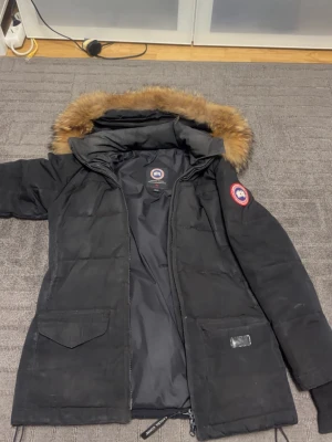 Svart Canada Goose parkas XS - Säljer en svart parkas från Canada Goose i storlek XS. Jackan har en stor huva med fluffig pälsdetalj, två stora fickor framtill och klassisk Canada Goose-logga på ärmen. Stängs med dragkedja och är fodrad med dun för extra värme.