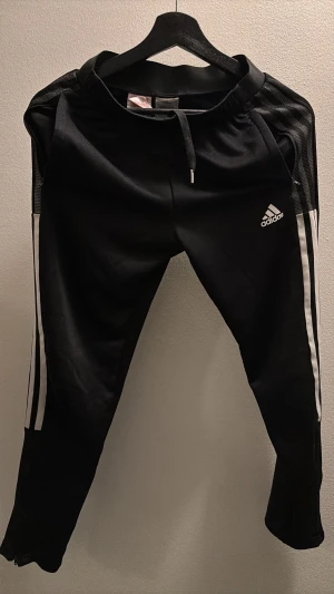 Svarta Adidas träningsbyxor med ränder - Säljer ett par svarta träningsbyxor från Adidas med vita ränder längs sidorna och logga på låret. Byxorna har snörning i midjan och är tillverkade i ett glansigt syntetmaterial. 