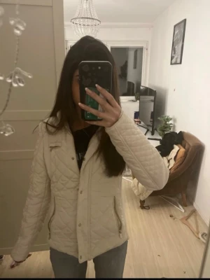 Vit quiltad jacka med dragkedjor - Säljer en beige quiltad jacka med snygga guldfärgade dragkedjor på fickorna. Jackan har en figurnära passform och är långärmad. Perfekt för dig som vill ha en stilren och enkel jacka till vardags.