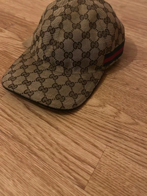 Gucci beige GG keps S - Säljer en beige keps från Gucci med klassiskt GG-monogram över hela kepsen och ikoniska gröna/röda ränder på sidan. Justerbart läderband bak och insida med svart tyg. Perfekt för dig som vill ha en lyxig och tidlös accessoar. Skicket är som nytt inte använd mycket.  