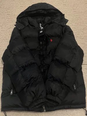 Svart pufferjacka från Polo Ralph Lauren - Säljer en svart pufferjacka från Polo Ralph Lauren med huva och dragkedjor. Jackan har det klassiska röda Polo-logot broderat på bröstet och flera praktiska fickor med dragkedja. Perfekt för kalla dagar och riktigt snygg till streetwear-stilen.