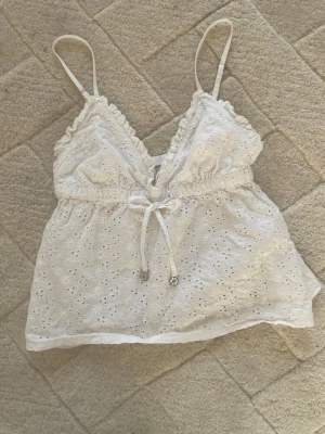 Vit broderad linnetopp från Censored - Supersöt vit linnetopp från Censored i storlek XS. Toppen har tunna axelband, broderade blommor över hela tyget och en söt knytning framtill. Volangkant vid bysten och lätt v-ringad. Perfekt för sommardagar och riktigt luftig känsla.