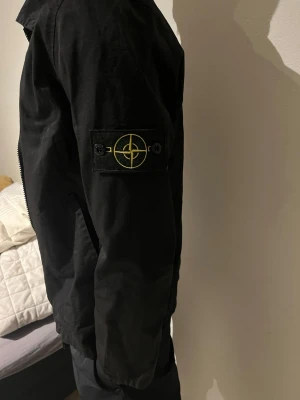 Svart vindjacka från Stone Island - Säljer en svart vindjacka från Stone Island med klassisk patch på vänster ärm. Jackan har dragkedja framtill, normal passform och är tillverkad i lätt polyester. Perfekt för dig som vill ha en clean och stilren look. Stl S/M