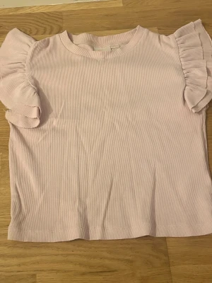 Ljusrosa ribbad topp med volangärm - Supersöt ljusrosa topp med ribbad struktur och volangdetaljer på ärmarna. Perfekt för dig som gillar en feminin och trendig stil. Toppen har rund halsringning och är i ett mjukt, stretchigt material.