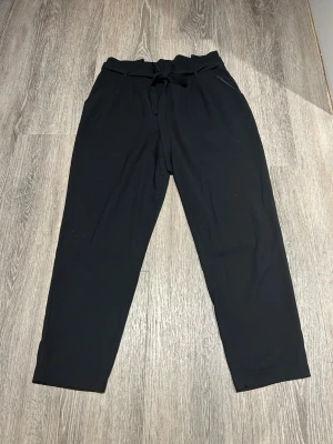 Kostymbyxor med knytband - Svarta kostymbyxor med hög midja och knytband i midjan. Byxorna har raka ben och bälteshällor, vilket ger en stilren och modern look. Perfekta för dig som gillar en clean och enkel stil. Strl. 40, men är lite små i strl så kanske 38.