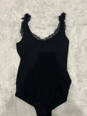 Svart spetsbody från Intimissimi - Säljer en svart body från Intimissimi med fina spetsdetaljer längs axelbanden och urringningen. Bodyn har en klassisk, enkel form och knäppning nedtill. Perfekt att styla till jeans eller kjol för en trendig look.