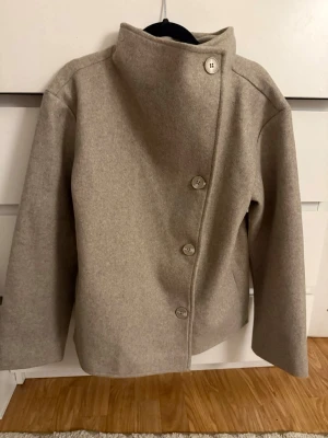 Beige kappa - Snygg beige ullrock med asymmetrisk knäppning och stora knappar. Liknar den virala kappan. Jackan har hög krage som kan bäras uppvikt eller nedvikt och är helfodrad för extra komfort. Perfekt för dig som gillar stilrena och minimalistiska plagg. Helt ny och oanvänd, stl 158/164 skulle säga xs/s❤️
