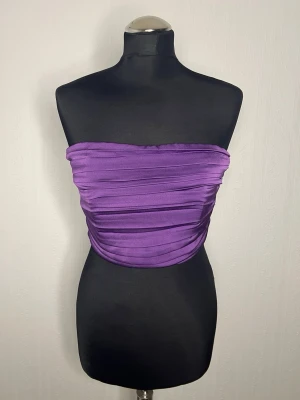 Lila bandeau tubtopp i satin - Säljer en snygg lila bandeau tubtopp i glansigt satinliknande material. Toppen har draperade detaljer som ger en cool och trendig look. Perfekt att styla med höga jeans eller kjol för en statement outfit.