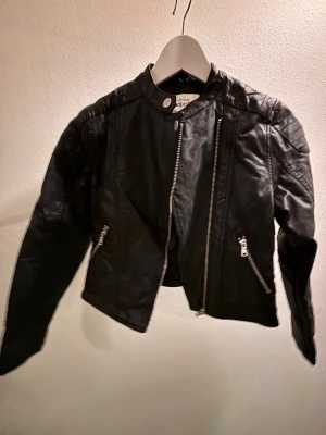 Svart jacka i skinnimitation med dragkedjor KappAhl - Säljer en svart jacka i skinnimitation från KappAhl med detaljer som snedställda dragkedjor och ståkrage med tryckknapp. Jackan har lång ärm och markerade axlar.