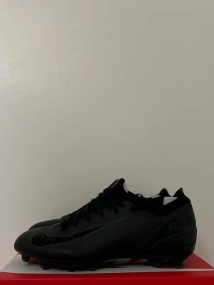 Svarta Nike fotbollsskor med dobbar - Säljer ett par helsvarta Nike fotbollsskor med klassisk låg profil och snörning. Skorna har en slimmad design, syntetmaterial och dobbar för bra grepp på planen. Swoosh-logga på sidan och diskreta detaljer för en clean look.