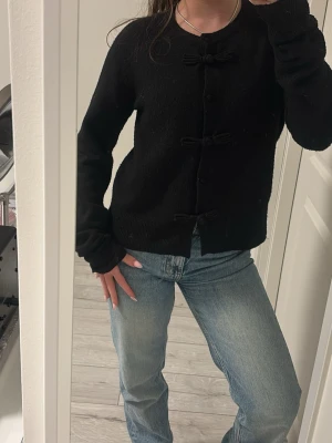 Svart kofta med rosetter - Säljer en svart kofta med långa ärmar och fina rosettdetaljer längs knäppningen framtill. Koftan är stickad och har en klassisk, enkel design som passar till mycket. Perfekt för dig som gillar stilrena plagg med en söt twist.