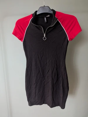 Svart och röd kortklänning Divided - Sportig kortklänning från Divided med svarta och röda partier samt vita detaljer längs ärmarna. Klänningen har korta ärmar, hög krage och en cool dragkedja med ring framtill. Materialet är mjukt och stretchigt, perfekt för en avslappnad look.