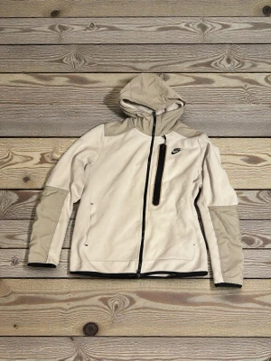Beige vindjacka från Nike med huva - Säljer en beige vindjacka från Nike med huva och svarta detaljer. Jackan har dragkedja framtill, en bröstficka med dragkedja och förstärkta partier på axlar och armbågar. Perfekt för dig som gillar sportig stil och vill ha något lätt och funktionellt.