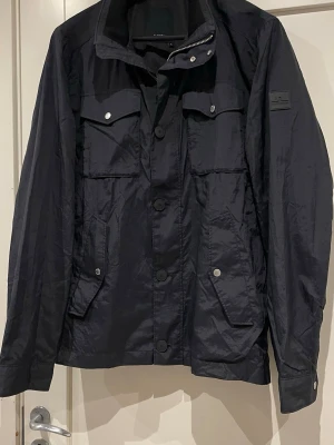 Svart vindjacka från J.Lindeberg L - Säljer en svart vindjacka från J.Lindeberg i storlek XL. Men sitter mer som en LJackan har flera fickor med knappar, hög krage och diskret logga på ärmen. Tillverkad i lätt polyester med knapp- och dragkedjestängning. Perfekt för dig som vill ha en clean och stilren look.