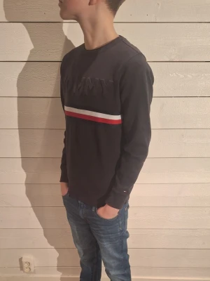 Mörkblå Tommy Hilfiger sweatshirt - Säljer en mörkblå sweatshirt från Tommy Hilfiger med upphöjd TOMMY-text på bröstet och en rand i rött, vitt och blått över bröstet. Tröjan har rund halsringning och långa ärmar. Perfekt för dig som gillar klassisk streetstyle.