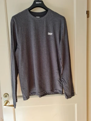 Grå långärmad tröja från DKNY strl XL - Snygg grå långärmad tröja från DKNY i storlek XL. Tröjan har en enkel och stilren design med liten vit DKNY-logga på bröstet. Tillverkad i 100% bomull. Aldrig använd.