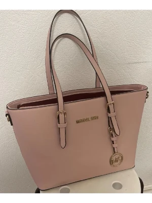 Rosa axelväska från Michael Kors - Snygg rosa axelväska från Michael Kors i skinn med guldfärgade detaljer och logotyp. Väskan har två långa handtag, dragkedja upptill och flera fack inuti med vinrött foder. Perfekt storlek för allt du behöver och stilren design med MK-hänge.