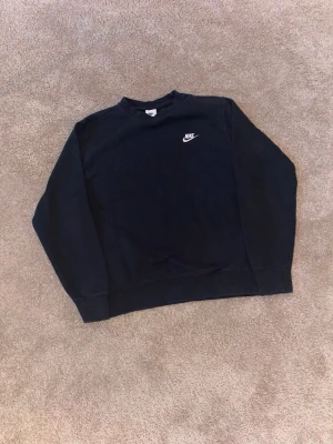 Svart Nike sweatshirt i bomull - Klassisk svart sweatshirt från Nike med broderad vit logga på bröstet. Tröjan har rund halsringning, långa ärmar och är gjord i mjuk bomull. Perfekt för dig som gillar enkel och stilren streetwear.