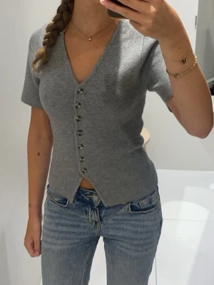 Grå kortärmad topp med knappar - Säljer en grå, kortärmad topp med v-ringning och dekorativa knappar framtill. Toppen har en figurnära passform och är tillverkad i ett mjukt stickat material. Knappen längst upp har lossnat. Bortsett från det så är den i bra skick.