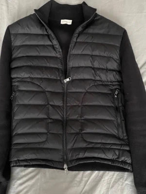 Svart dunväst från Moncler - Säljer en svart dunväst från Moncler med quiltad framsida och dragkedja. Västen har en stilren look med Moncler-logga på ärmen och ribbade detaljer. Perfekt för lager-på-lager och snygg till både jeans och hoodie.