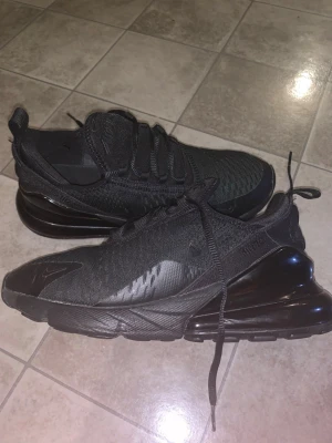 Svarta Nike Air Max 270 sneakers - Säljer ett par helsvarta Nike Air Max 270 sneakers med stor synlig luftbubbla i hälen. Ovandelen är i mesh och syntet, med diskreta Nike-loggor på sidan och tungan. Skorna har snörning och en chunky, sportig vibe som passar perfekt till streetwear.